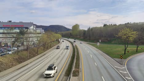 Vista-Aérea-Con-Vistas-A-La-Autopista-E39-De-Cuatro-Carriles-En-Asane,-Con-Centros-Comerciales,-Montañas-Y-Tráfico-En-Dirección-A-Bergen.