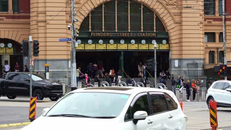 Vehicles-traffic-on-Swanston-and-Flinders-streets-with-iconic-Flinders-Street-Station-entrance-as-backdrop,-creating-a-vibrant-transit-hub-for-commuters-and-tourists,-and-a-bustling-urban-scene