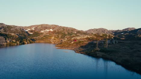 Un-Hombre-Sentado-Junto-A-Su-Tienda-Roja-En-El-Paisaje-Accidentado-Que-Rodea-El-Lago-En-Skurven,-Eidsvoll,-Akershus,-Noruega---Drone-Volando-Hacia-Adelante