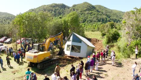 Aerial-View-Of-Excavator-Pulling-House-Across-River-In-Chiloe-With-Local-Community-Watching