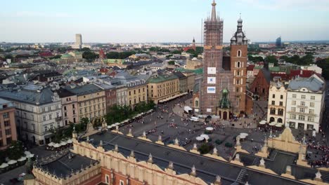 Toma-De-Drones-De-La-Plaza-Rynek-Glowny,-Basílica-De-La-Construcción-De-Restauración-De-La-Torre-De-La-Iglesia-De-Santa-María-En-La-Plaza-Principal-Del-Mercado-De-Cracovia-Rodeada-De-Edificios