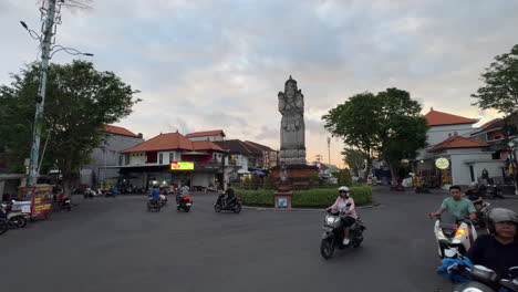 A-roundabout-in-Bali-with-a-tall-statue-in-the-center,-surrounded-by-motorbikes,-buildings,-and-trees-at-dusk