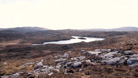 Loch-of-water-nestled-amongst-rugged-mountainous-terrain-and-golden-brown-tussocks-with-boulders-in-Outer-Hebrides-of-Scotland-UK