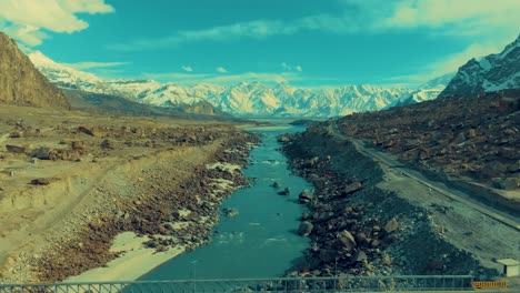 Tilt-up-drone-shot-of-snow-covered-ranges-at-background-with-beautiful-landscape-of-canal-leading-to-mountains-in-Skardu,-Pakistan