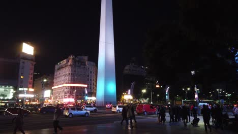 An-evening-at-the-obelisk-of-Buenos-Aires-City,-people-walk-in-autumn-night-with-traffic-and-neon-signs