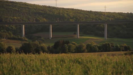 High-speed-train-crossing-a-lush-green-landscape-on-a-bridge-at-sunset,-wind-turbines-in-the-background