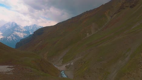 4K-Drohnenaufnahme-Mit-Vorwärtsflug-über-Grasbedeckte-Berge-Mit-Schneebedeckten-Bergen-Und-Dunklen-Wolken-Im-Hintergrund