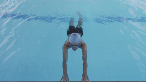 Un-Joven-Nadador-Usa-Gafas-Y-Gorra-Entrenando-El-Movimiento-De-Las-Piernas-En-El-Agua-Mientras-Sostiene-El-Costado-De-La-Piscina.