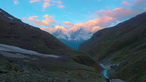 4K-Drohnenaufnahme-Einer-Schneebedeckten-Himalaya-Bergkette-Mit-Goldenem-Sonnenlicht-Und-Einem-Fließenden-Gletscherfluss