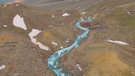 4K-Drohnenaufnahme-Mit-Schwenkfunktion-Eines-Flusses,-Der-Aus-Einem-Gletschersee-Im-Himalaya-Entspringt-Und-Schneebedeckte-Berge-Freigibt