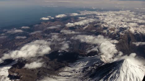 Vista-Aérea-Desde-El-Avión-De-Nubes-Y-Paisaje-Montañoso-De-Irán-Cubierto-De-Nieve-En-Medio-Oriente