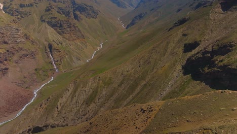 4K-Drone-shot-revealing-a-gorge-with-a-stream-running-through-it-with-waterfalls-and-snow-capped-mountains