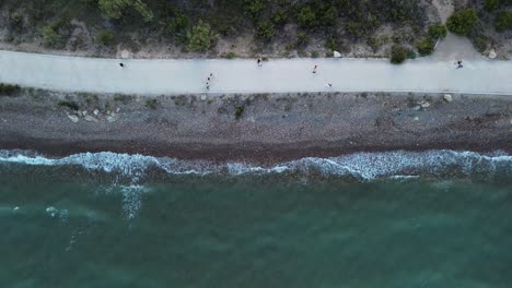 Drone's-top-down-view-of-a-serene-beach-with-waves-gently-hitting-the-shore-at-sunset,-as-people-walk-and-exercise-along-the-shoreline