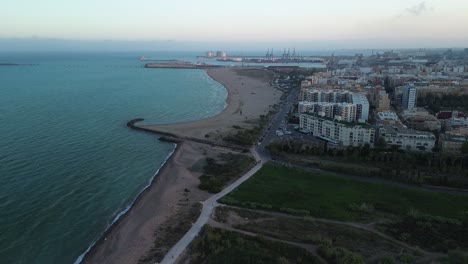 Seitliche-Luftaufnahme-Eines-Stadtgebäudes-Am-Strand-Mit-Dem-Seehafen-Im-Hintergrund