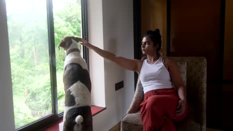 Woman-in-white-tank-top-pets-dog-while-sitting-by-window,-looking-out-at-lush-green-scenery