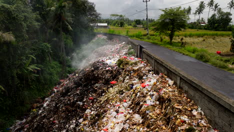 Smoldering-garbage-on-roadside-of-Bali-countryside-road,-low-aerial-dolly