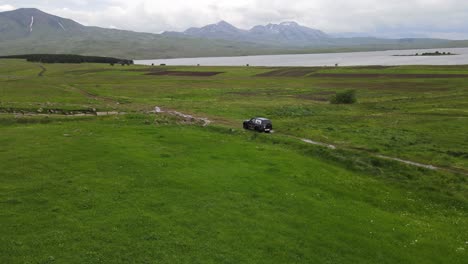 A-car-is-parked-on-a-lush-green-field-by-a-serene-lake,-surrounded-by-rolling-hills-and-mountains-in-the-distance