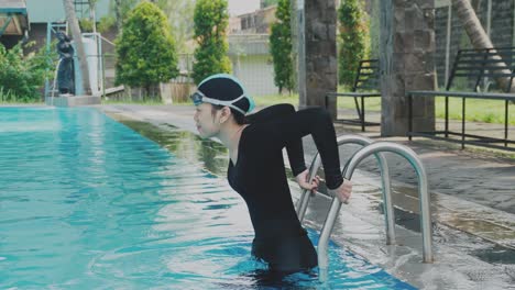 Young-Female-Swimmer-In-A-Black-Swimsuit-Enters-The-Pool-On-The-Steps-And-Wearing-Swim-Goggles