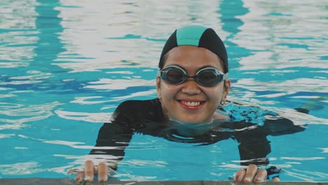 Una-Joven-Asiática-Usa-Gorra-Y-Gafas-Aprendiendo-A-Nadar-Sosteniendo-El-Lado-De-La-Piscina-Y-Moviendo-Las-Piernas-Bajo-El-Agua.