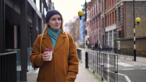 Happy-cheerful-positive-and-relaxed-young-Caucasian-woman-enjoying-the-weather,-walking-down-a-London-street-with-a-coffee-cup-in-her-hands,-and-smiling-while-looking-around