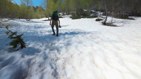 Hiker-with-backpack-and-camera-walks-up-a-snowy-mountain-slope-with-trees