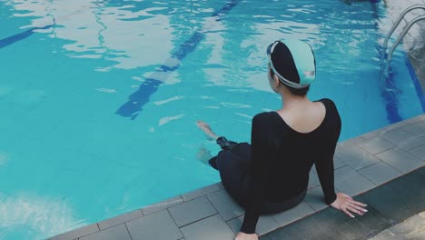 Rear-View-Of-Young-Woman-Swimmer-Sitting-On-The-Poolside-With-Legs-In-Blue-Water