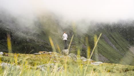 Man-standing-on-small-rock-at-the-edge-of-a-cliff,-enjoying-cloudy-view-of-mountains,-filming-the-scenery-and-taking-in-the-moment