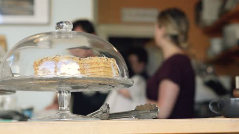 A-blurred-shot-of-co-workers-talking,-with-a-cake-in-a-glass-dome-in-the-foreground,-in-a-local-coffee-shop-|-Gourmet-Coffee-House-|-Shot-in-HD-at-cinematic-24-fps