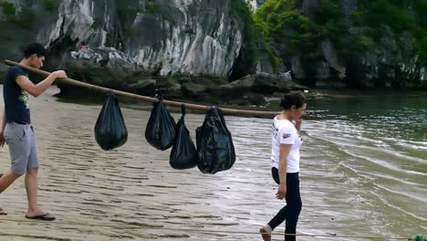 Ha-Long-Bay,-Vietnam---A-man-and-woman-carrying-heavy-loads-using-a-long-bamboo-stick-in-tandem---wide-shot