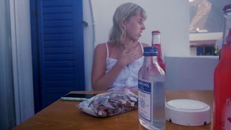 Young-Woman-Sitting-Down-To-Hear-Seat-By-The-Balcony-in-The-Warm-Summer-Evening-In-Santorini-Greece-Where-People-Have-a-Pre-Party-Before-Going-Out