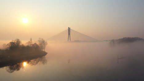 Low-flying-towards-over-a-river,-towards-a-modern-cable-bridge-during-a-foggy-sunrise-morning