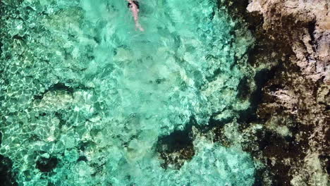 Top-down-drone-shot-of-a-young,-caucasian-man-swimming-in-crystal-clear-water-on-comino-island