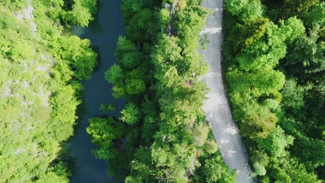 Aerial-top-view-of-a-river-flowing-next-to-a-country-road-and-surrounded-by-a-green-mountain-forest