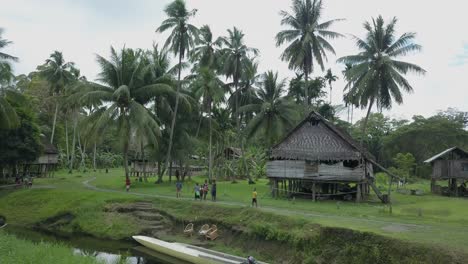 Aerial-view-slow-moving-shot,-kids-standing-near-the-river,-huts-and-palm-trees-in-the-background