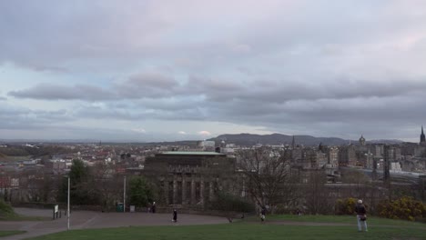 Panning-shot-from-Calton-hill-with-people-walking-and-Nelson-monument-in-the-foreground-with-nice-sunset-light-and-clouds-overlooking-the-city-of-Edinburgh,-Scotland