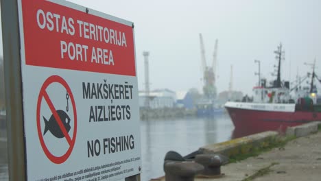 No-fishing-at-port-area-sign-at-Port-of-Liepaja-with-out-of-focus-fisherman-ships-and-port-cranes-in-background,-foggy-day,-close-up