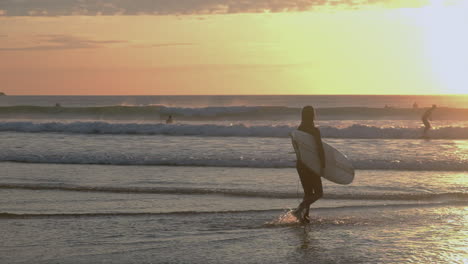 Young-female-walking-into-the-sea-carrying-a-surfboard-at-sunset