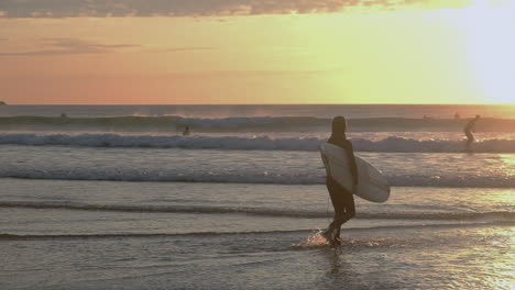 Mujer-Joven-Caminando-Hacia-El-Mar-Llevando-Una-Tabla-De-Surf-Al-Atardecer