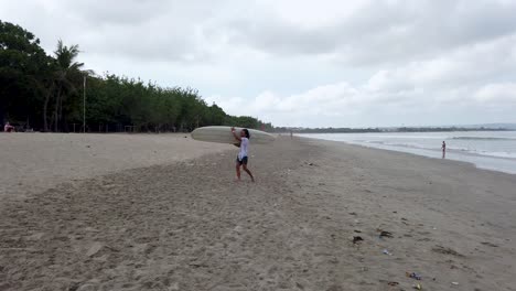 Mann,-Der-Mit-Einem-Surfbrett-Am-Weißen-Sandstrand-Von-Kuta,-Bali,-Asien,-Spaziert,-An-Bewölkten-Tagen-Inmitten-Der-Reisebeschränkungen-Des-Coronavirus-Covid-19