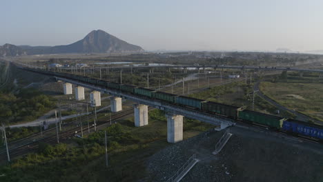 Full-loaded-coal-cargo-carriages-train-stopped-on-a-railway-bridge-with-road-and-mountain-ridge-in-the-background,-on-the-sunset