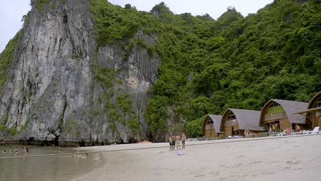 Tourists-Enjoying-A-Walk-On-The-Sandy-Beach-Of-Ha-Long-Bay,-Vietnam-With-The-Beautiful-Limestone-Rock-Formation-In-The-Background---Wide-Pan