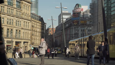 4K,-Shoppers-and-commuters-busy-themselves-in-Exchange-Square,-Manchester-City,-tram-runs-along-track-to-right-hand-side-of-shot