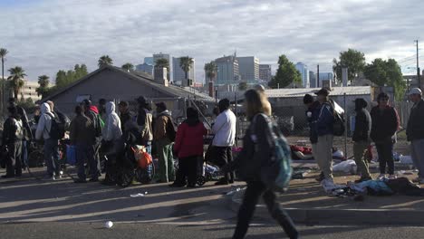 Un-Ciclista-Pasa-Junto-A-Una-Larga-Fila-De-Personas-Sin-Hogar-Que-Esperan-Conseguir-Artículos-De-Primera-Necesidad-Para-Sobrevivir-En-Las-Calles-Urbanas-De-Phoenix,-Arizona.