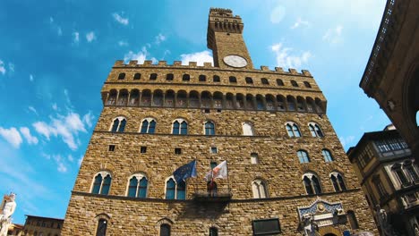 Old-Palace-in-Florence-with-waving-flags-on-its-balcony-and-blue-sky-above,-wide-angle