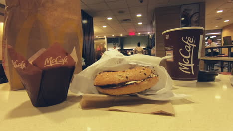 Editorial,-view-of-McDonald's-breakfast-meal,-coffee,-bagel-sandwich-and-muffin-on-fast-food-restaurant-table,-and-people,-clients-passing