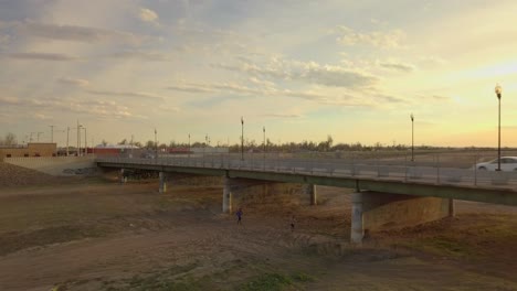 Two-people-running-on-dry-river-bed-under-bridge-at-sunset,-aerial-rising-pull-back