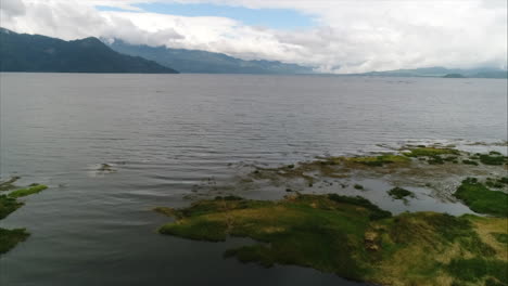 AERIAL:-Panoramic-shot-of-lake-shore-sorrounded-by-mountains-and-blue-sky,-Yojoa,-Honduras