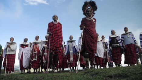 Eine-Gruppe-Von-Dorfbewohnern-Des-Massai-Stammes-Tanzt-Und-Springt-Hoch-Für-Eine-Kulturelle-Traditionelle-Zeremonie-In-Ihrer-Traditionellen-Kleidung-Im-Wüstenbusch-Von-Tansania,-Afrika---Zeitlupen-Gänseblümchenaufnahme