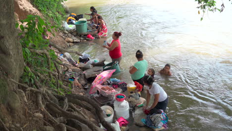 Mehrere-Frauen-Waschen-Ihre-Kleidung-Am-Ufer-Des-örtlichen-Flusses,-Während-Ihre-Kinder-In-Der-Nähe-Schwimmen-Und-Spielen