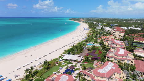 Zoom-in-aerial-4K-footage-revealing-the-sunlit-Grace-Bay-coastline,-white-sandy-beach,-turquoise-waters,-and-green-beach-resorts-with-rows-of-sun-beds-and-umbrellas
